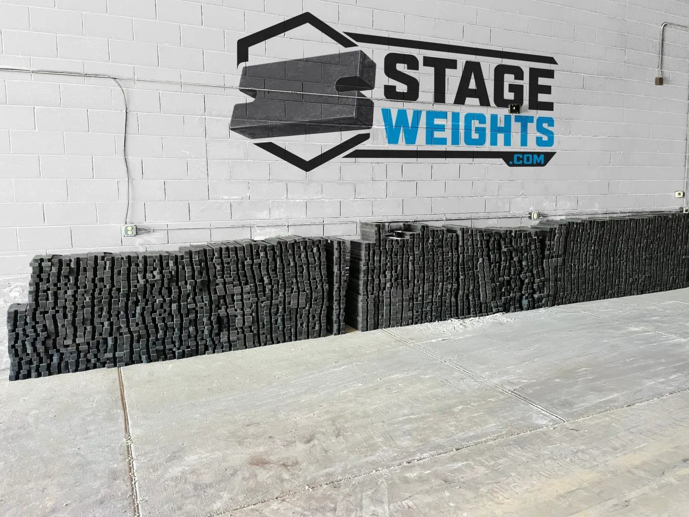 Long rows of black steel stage weights stacked against a white warehouse wall painted with the Stage Weights logo